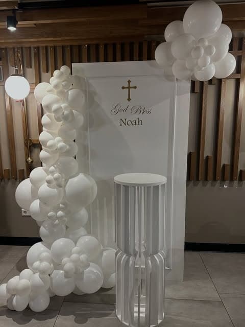 White christening backdrop reading God Bless Noah with a slim gold cross, white balloon garland and fluted pillar