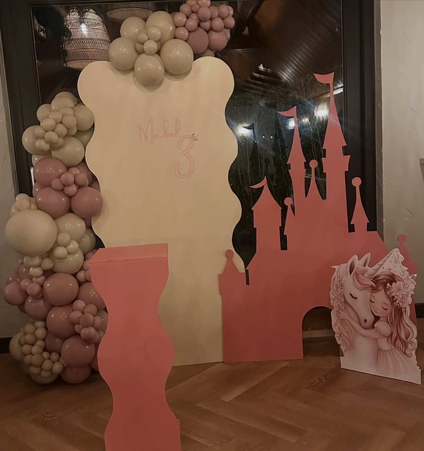 Third birthday princess castle set with a pink castle silhouette, cream wavy backdrop and dusty rose balloon garland, styled at night
