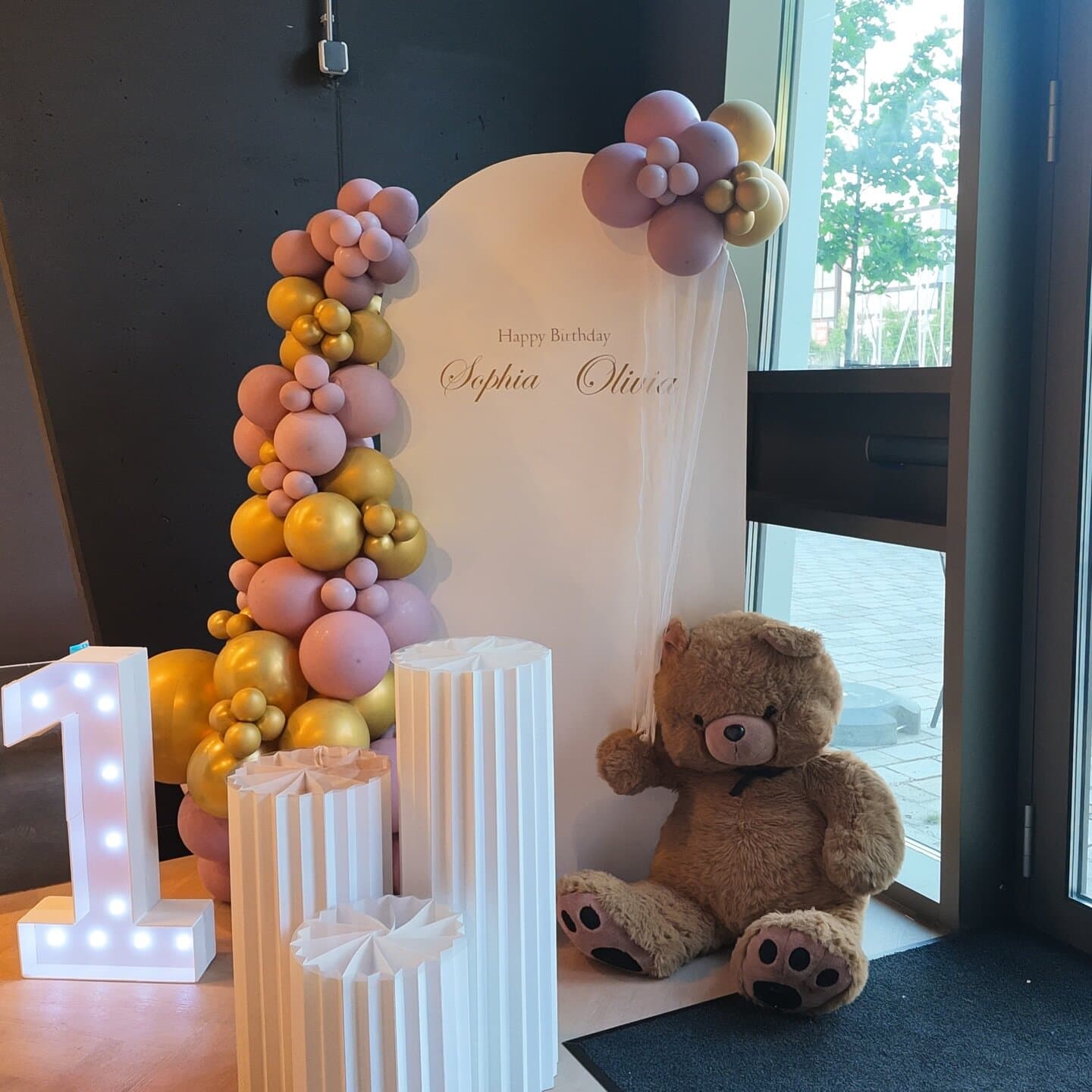 First birthday board for twins Sophia and Olivia with pink and gold balloons, a lit number one and a teddy bear
