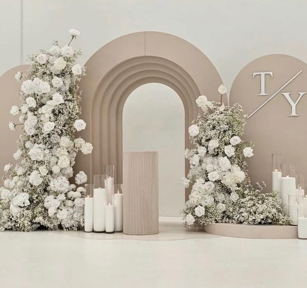 Sculptural taupe double wedding arch with white roses, baby's breath and pillar candles, finished with the couple's monogram
